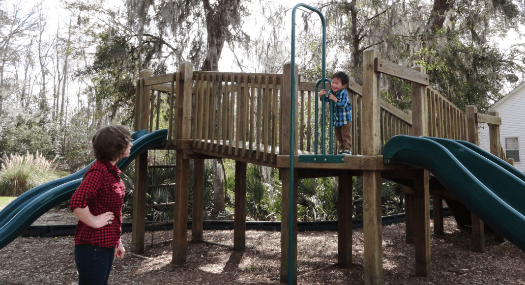 Adoption: A mother looks on at her smiling toddler son as he climbs playground equipment.