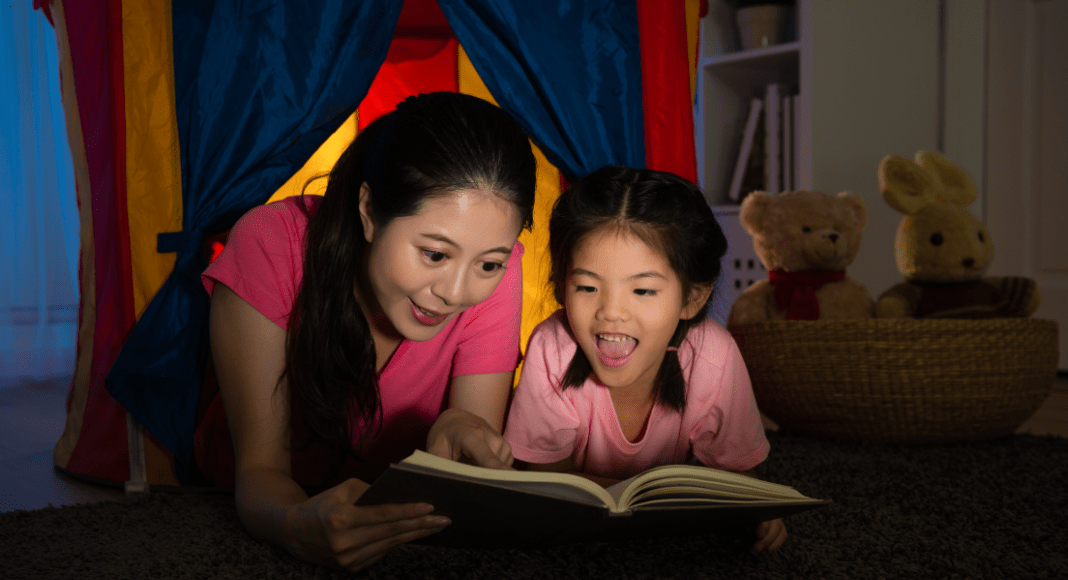 A mom reads a book with her daughter in a blanket fort.