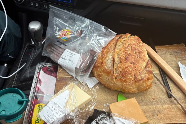 A round loaf of artisan bread, wedges of cheese, and packaged raspberries and roast beef are arranged on a wooden cutting board inside a parked car.
