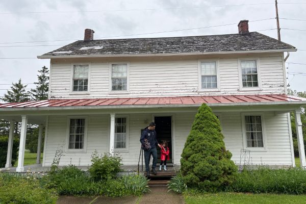 A father and daughter exit the historic Harriet Tubman Home for the Aged, a preserved white wooden home with an old roof, greenery in the front, and porch.